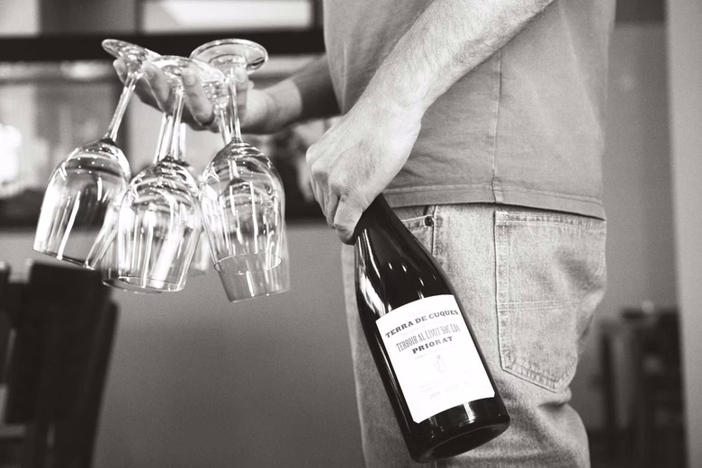 Person holding a bottle of wine and four upside-down wine glasses in one hand