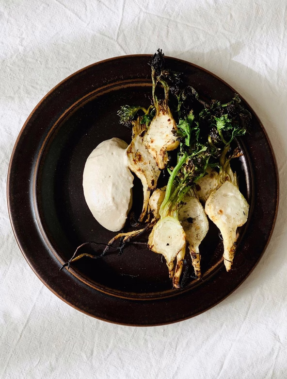 Grilled root vegetables with charred greens and a dollop of creamy sauce on a dark plate.