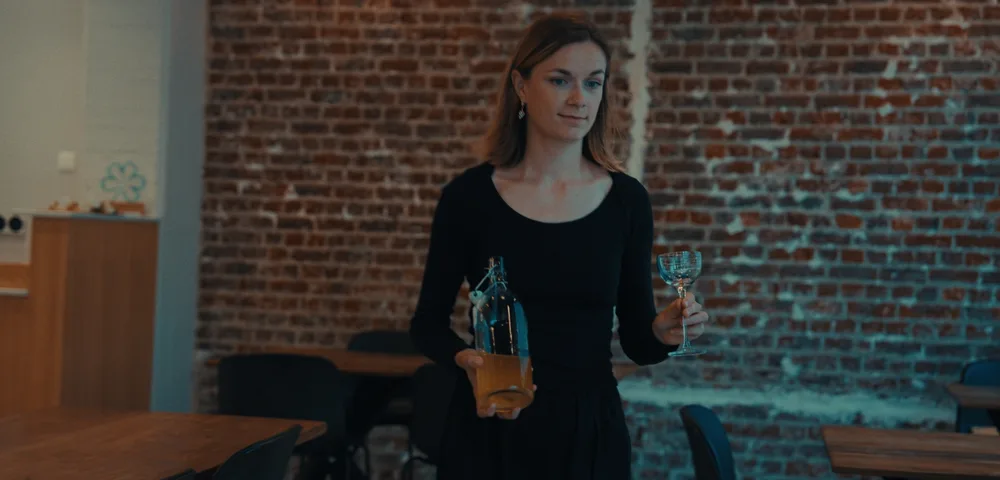 A woman in a black long-sleeved top and skirt walks through a restaurant with exposed brick walls, holding a swing-top bottle of amber liquid in one hand and an empty stemmed glass in the other. Wooden tables and dark chairs surround her.