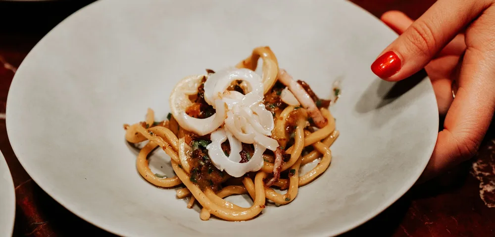 Handen met rode nagels plaatsen een wit diep bord met spaghetti en inktvisringen op een marmeren tafel. Op de achtergrond staan gelijkaardige borden klaar.