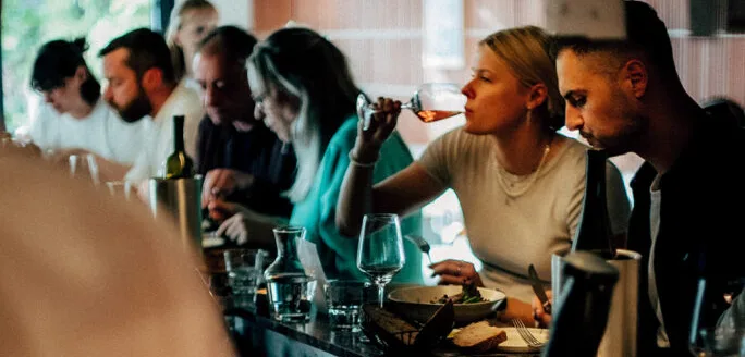 Gasten zitten aan een restaurantbar en eten en drinken. Een vrouw drinkt witte wijn. Op de bar staan borden, glazen en sauzen. Een kok staat op de voorgrond bij het aanrecht.