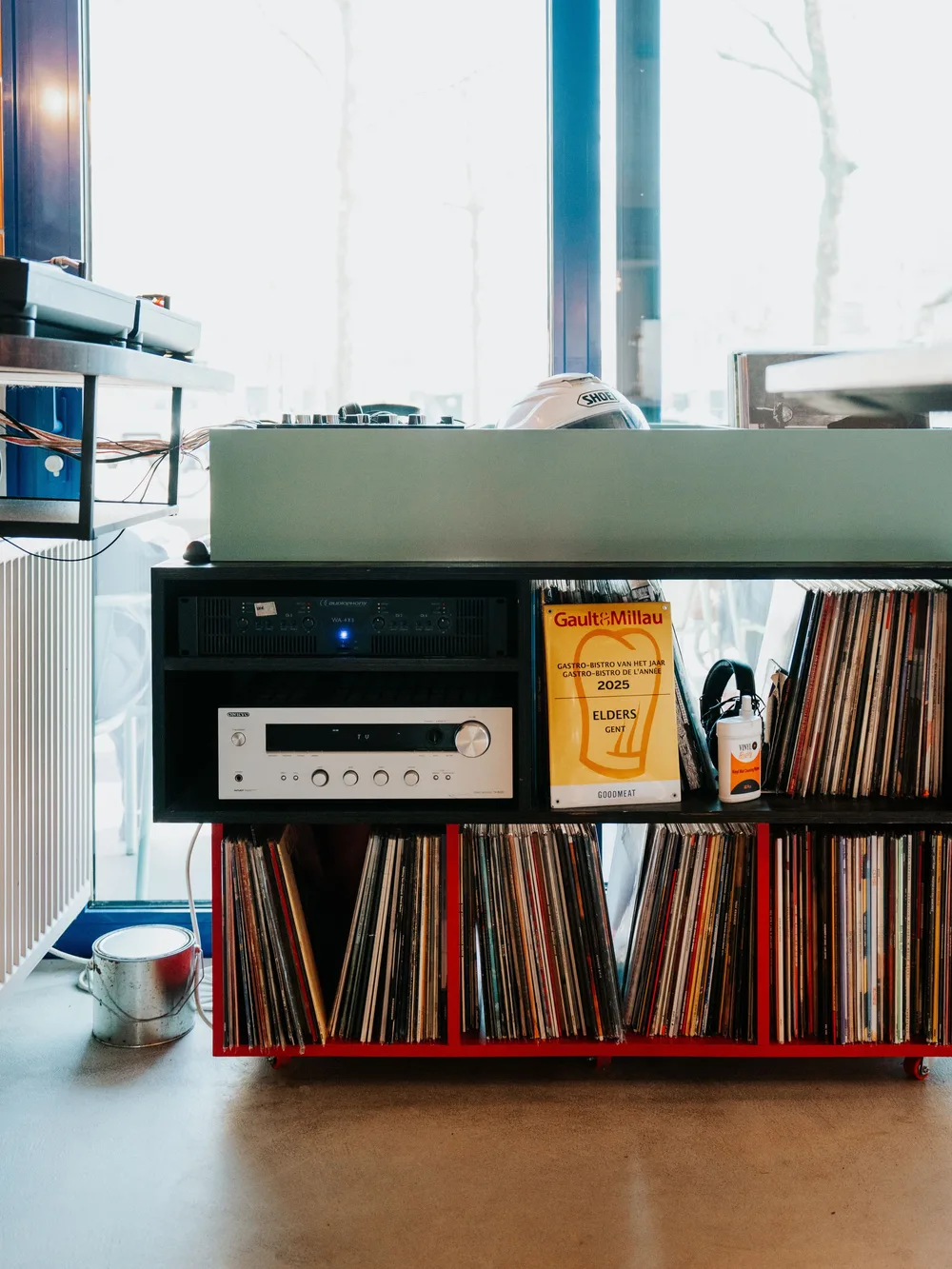 Een houten rek op wieltjes met vinylplaten, een versterker en een Gault&Millau 2025-award voor Elders Gent. Op het werkblad staat audioapparatuur. Op de achtergrond grote ramen met bomen.