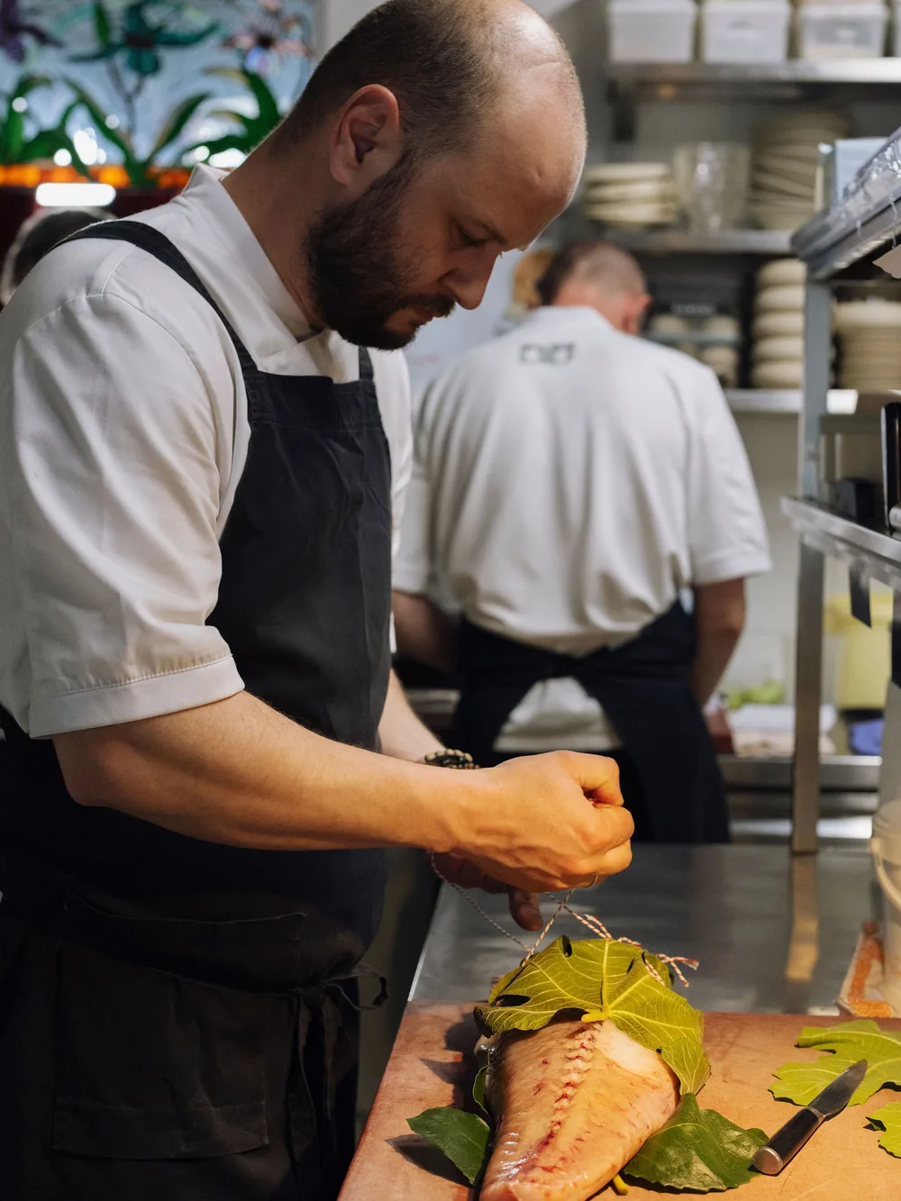 Chef in white shirt and dark apron ties twine around a fish fillet wrapped in large green leaves on a wooden board in a busy restaurant kitchen. A second chef works in the background.