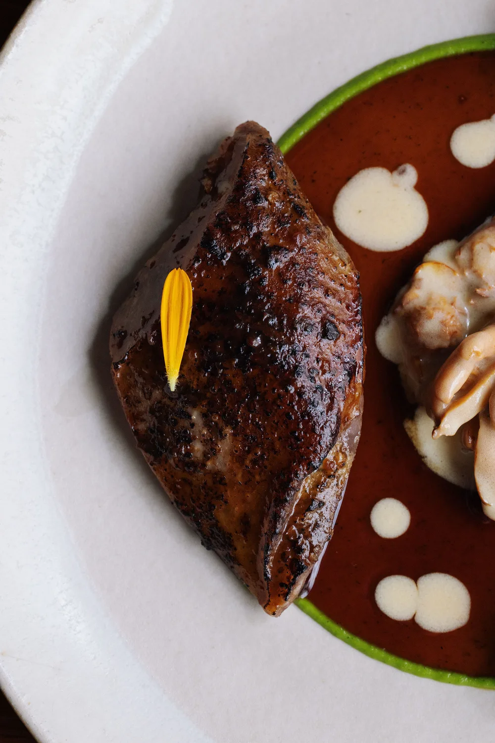A seared meat portion garnished with a yellow flower petal, plated on a white dish with a green rim, rich brown sauce, cream dots, and sautéed mushrooms alongside.