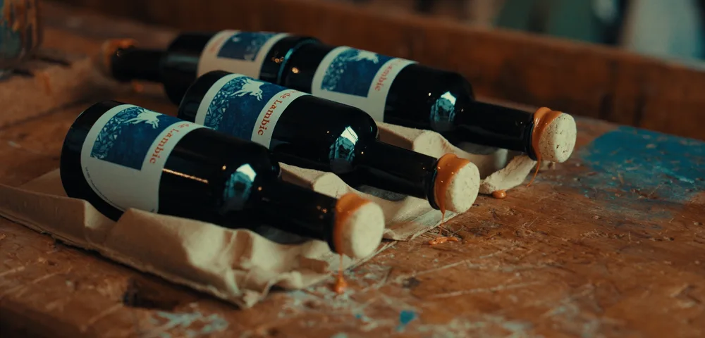 Quatre bouteilles de lambic couchées sur un support en carton, posées sur une table en bois. Les bouchons en liège laissent couler un liquide ambré. Étiquettes bleues et blanches visibles.