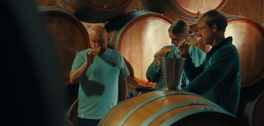 Trois personnes dégustent du vin blanc dans des verres à pied dans une cave, entourées de fûts en chêne empilés. Un seau en inox est posé sur un tonneau au premier plan.