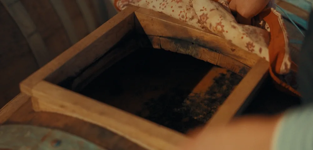 Mains tenant un linge fleuri au-dessus d'un cadre en bois carré posé sur un tonneau, contenant un liquide sombre, dans une ambiance chaleureuse tamisée.
