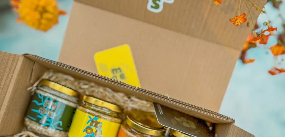 Open kartonnen doos met het logo "moes" op het deksel, met vier glazen potjes met gouden deksels op stro: donkergroen, geel, oranje en groen etiket. Op de achtergrond oranje en gele bloemen, onderaan een stuk jeansstof.