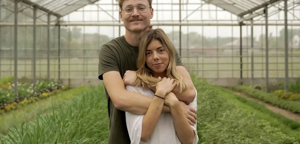 Een man met bril en een vrouw met blond haar staan innig omarmd in een grote glazen serre. Hij staat achter haar met zijn armen om haar heen. Rondom hen groeien rijen groene planten en kruiden, met bloemen links.