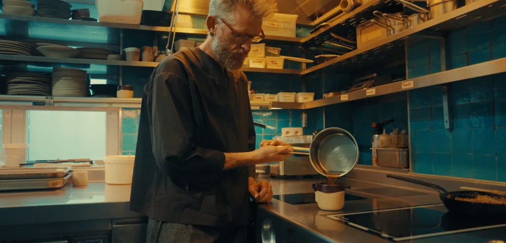 Een bebaarde man met grijs stekelhaar en bril staat in een professionele keuken met blauwe tegels en schenkt vloeistof uit een steelpan in een kleine witte kom. Rondom zijn rekken met borden, bakjes en potten.