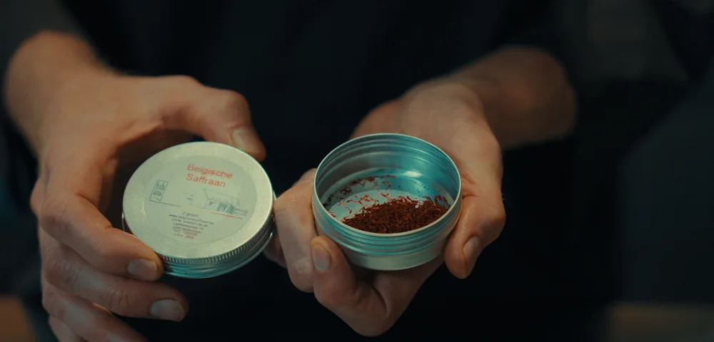 Twee handen houden een open aluminium blikje met rode saffraandraadjes vast, en in de andere hand het deksel met het opschrift "Belgische Saffraan" en "2 gram". Op de achtergrond is een donkere trui te zien.