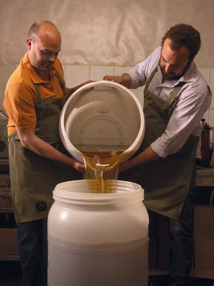 Deux hommes en tablier versent un liquide doré, probablement du miel ou de l'huile, d'un grand seau blanc dans un bidon dans un atelier alimentaire.