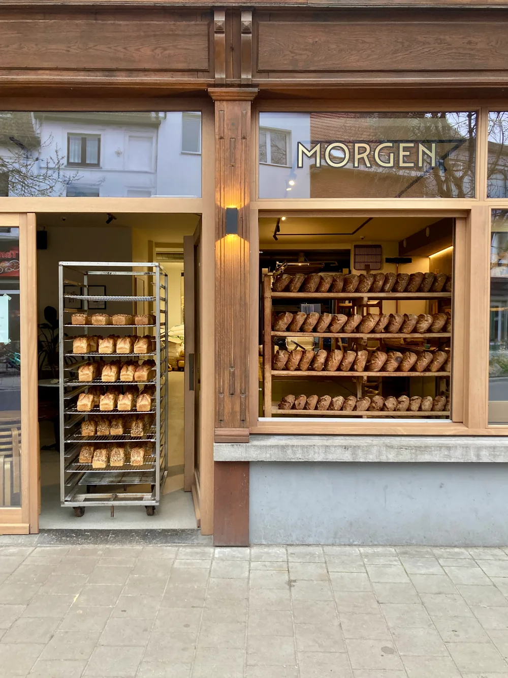 Exterior of Morgen bakery with wooden shopfront. A rack of golden loaf breads stands in the open doorway. Display shelves inside the window are lined with rows of rustic sourdough loaves.