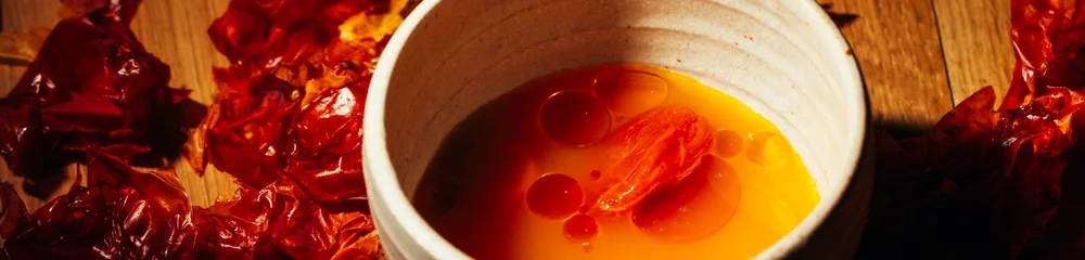Bol en céramique blanche contenant une sauce orange vif avec des filets rouges, entouré de piments séchés étalés sur une surface en bois, éclairé par une lumière chaude et directionnelle.