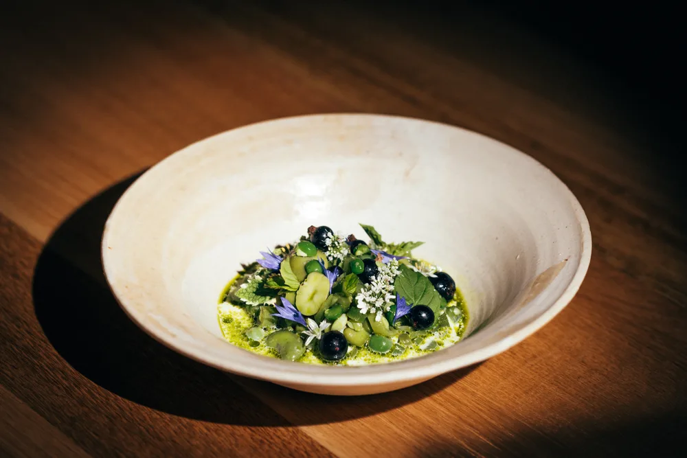 Bol en céramique blanche posé sur une table en bois, garni d'herbes fraîches, de petits pois, de baies noires, de fleurs de bourrache bleues et blanches, nappé d'un bouillon vert vif.