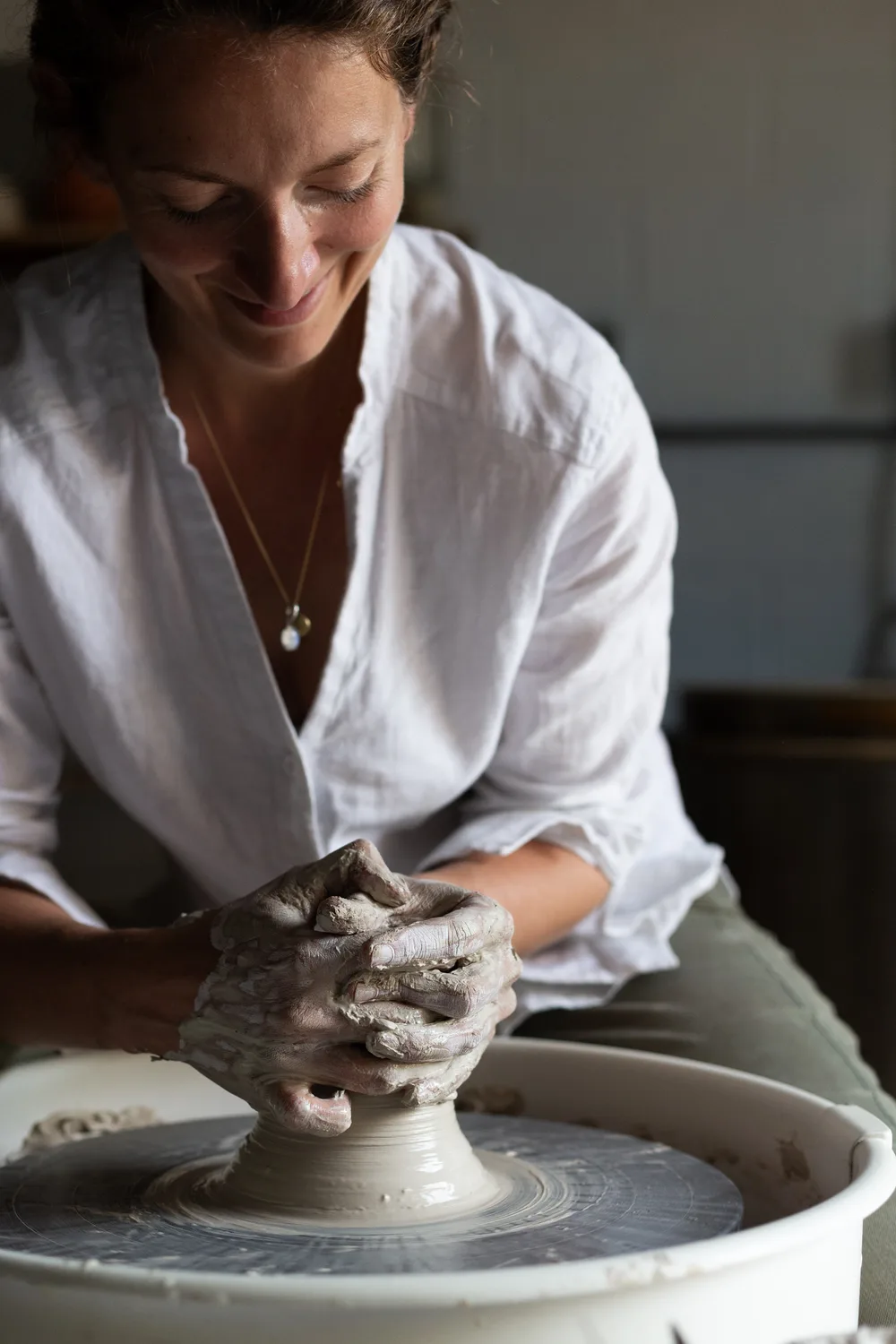 Une femme souriante en chemise blanche façonne de l'argile sur un tour de potier. Ses mains sont couvertes d'argile. Ambiance lumineuse et artisanale dans un atelier.
