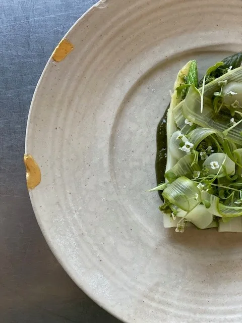 Assiette en céramique beige avec une composition de légumes verts — feuilles de chicorée, jeunes pousses et petites fleurs blanches — posée sur une surface en métal gris.