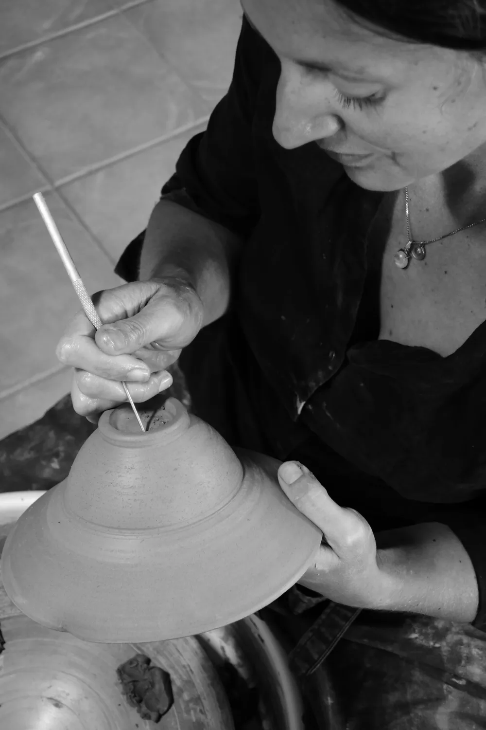 Femme en noir travaillant un bol en argile cru sur un tour de potier, tenant un outil de sculpture. Photo en noir et blanc, prise en plongée.