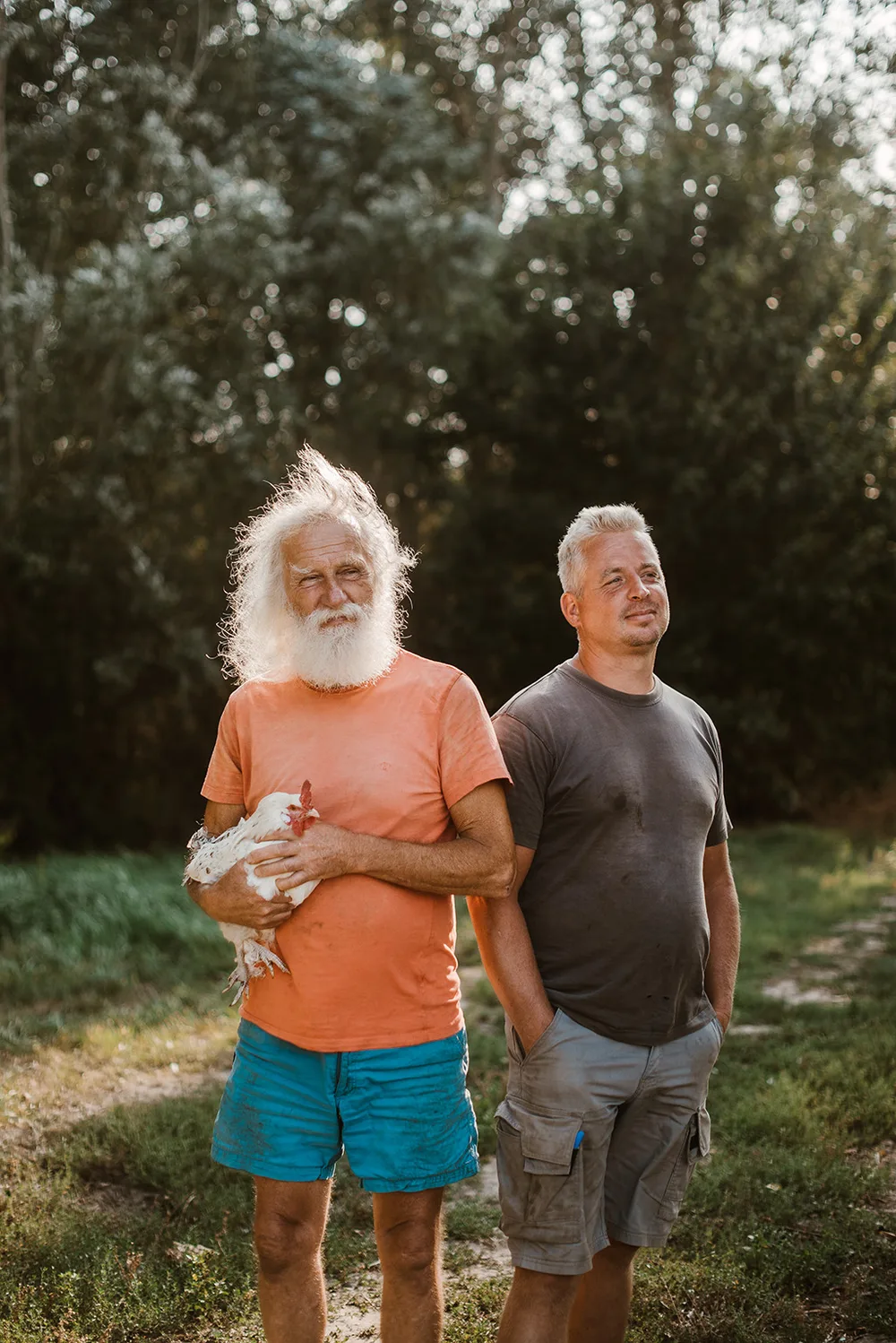 Twee mannen staan buiten op een grasveld met bomen op de achtergrond. De oudere man met wit haar en baard draagt een oranje T-shirt en houdt een witte kip vast.