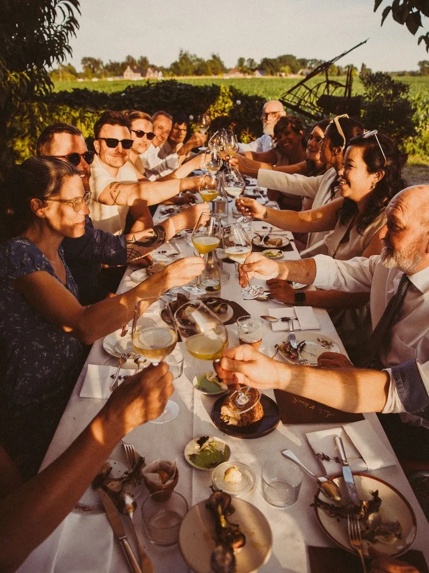 Groep mensen aan lange gedekte tafel buiten, die glazen heffen voor een toast. Zomerse sfeer, groene achtergrond. Tafel met borden, bestek en restjes eten. Gouden avondlicht.