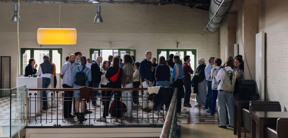 Een groep mensen staat te praten op een mezzanine in een industrieel pand met bakstenen muren, stalen dakconstructie, hangende lampen en groene raamkozijnen.