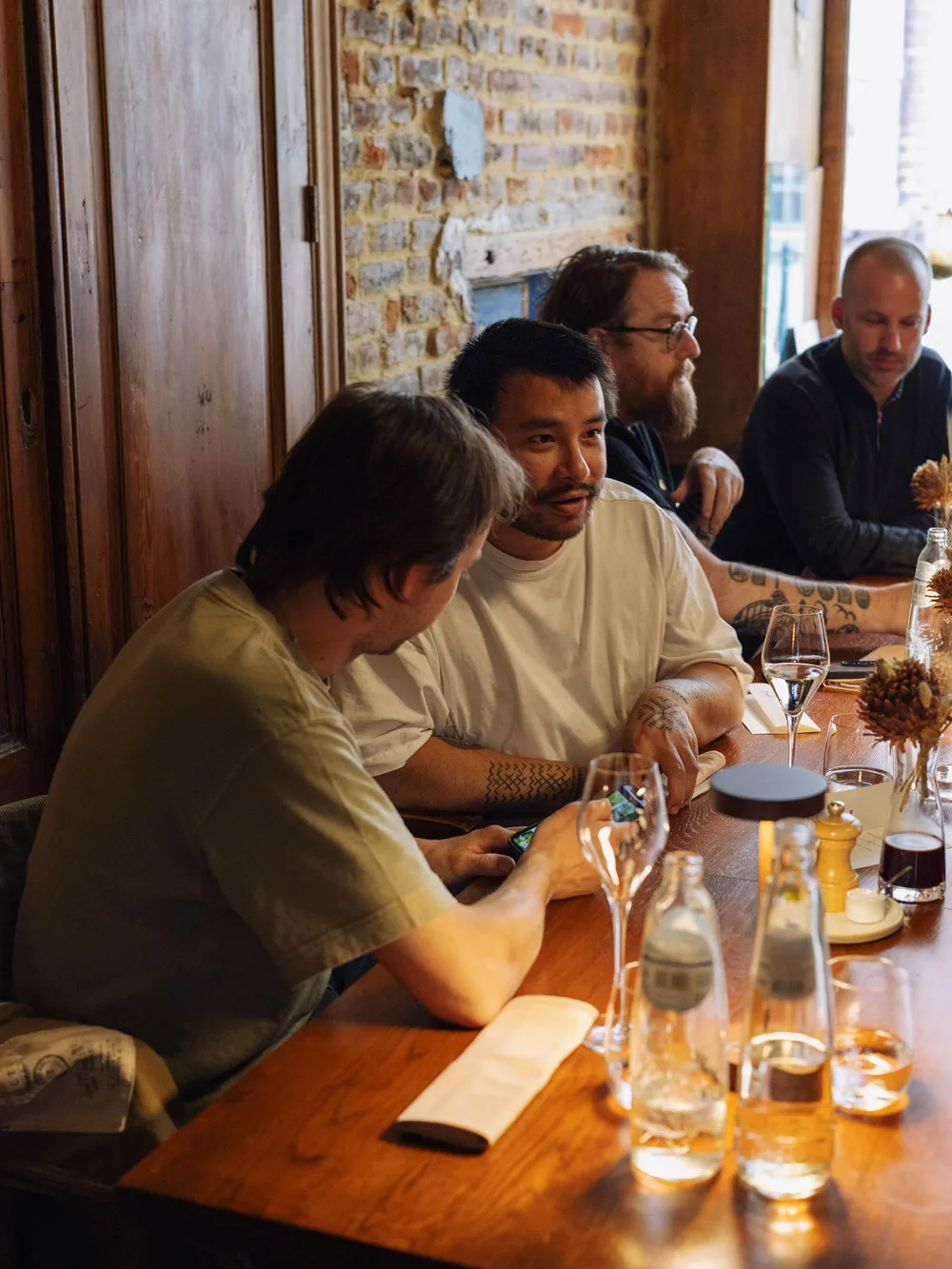 Vier mannen zitten aan een houten tafel in een restaurant met bakstenen muur. Op tafel staan glazen, waterflessen en kleine decoraties. De mannen zijn in gesprek.