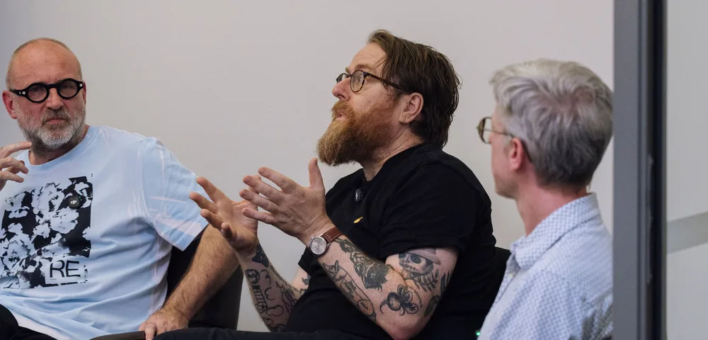 Drie mannen zitten in bruine stoelen in gesprek. De man in het midden, met baard, bril en tatoeages, gebaart met zijn handen. De anderen luisteren aandachtig. Setting is een moderne, witte ruimte.