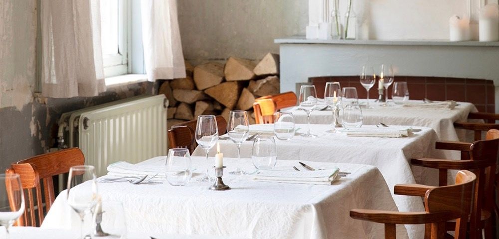 Dining tables with white tablecloths and wooden chairs in a softly lit rustic room with a window and floral decor.